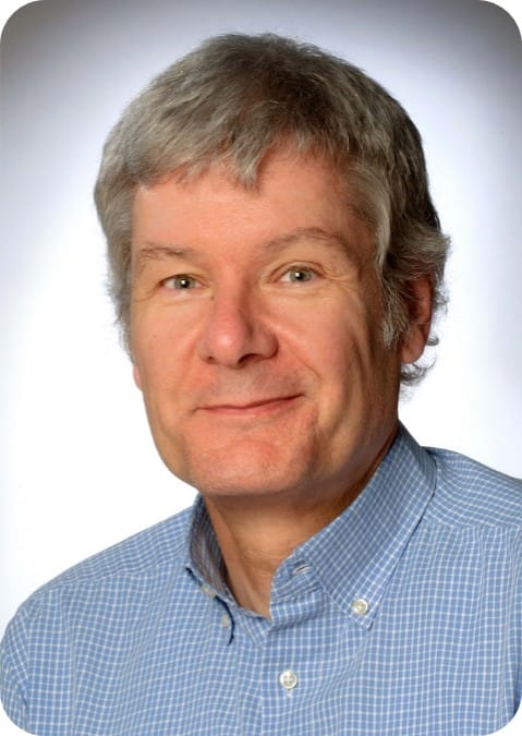 A middle-aged man with grey hair, wearing a blue checked shirt, faces the camera and smiles against a neutral background—an advocate for climate-resilient agriculture and advances in ecophysiology.