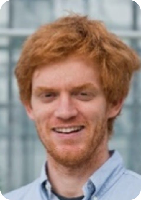 A man with red hair and a beard, wearing a light blue collared shirt, stands in front of a blurred background, representing MultiStress Research focused on food security and resilient crops like maize.