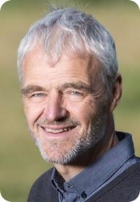 An older man with short grey hair and a beard is smiling outdoors, wearing a dark collared shirt and jumper. The blurred greenery hints at his work in Ecophysiology and MultiStress Research, possibly related to maize.