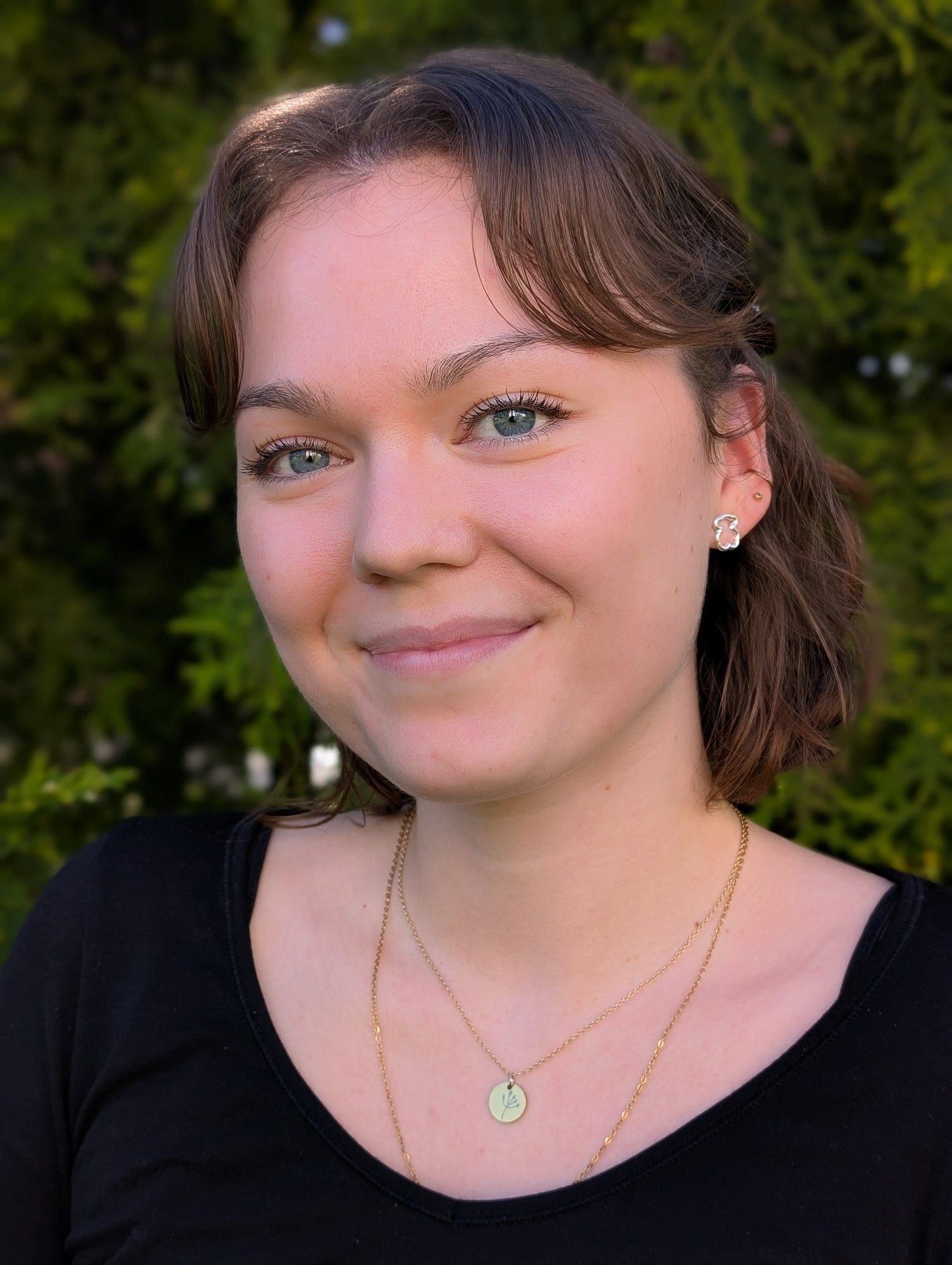 A young woman with short brown hair, wearing a black top and gold necklaces, smiles outdoors with green foliage and maize plants in the background.