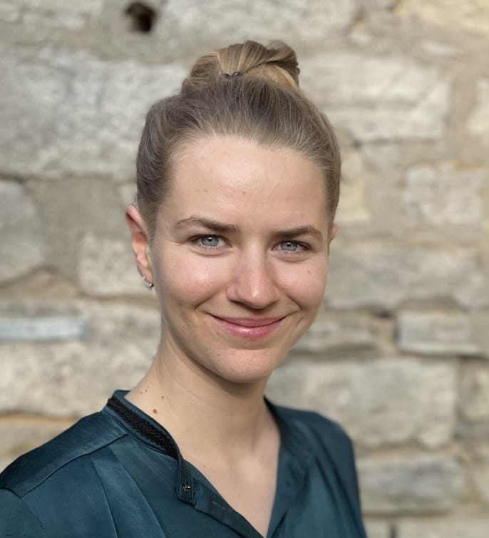 A woman with blonde hair tied in a bun, wearing a dark green top, smiles whilst standing in front of a stone wall, embodying the spirit of climate-resilient agriculture.