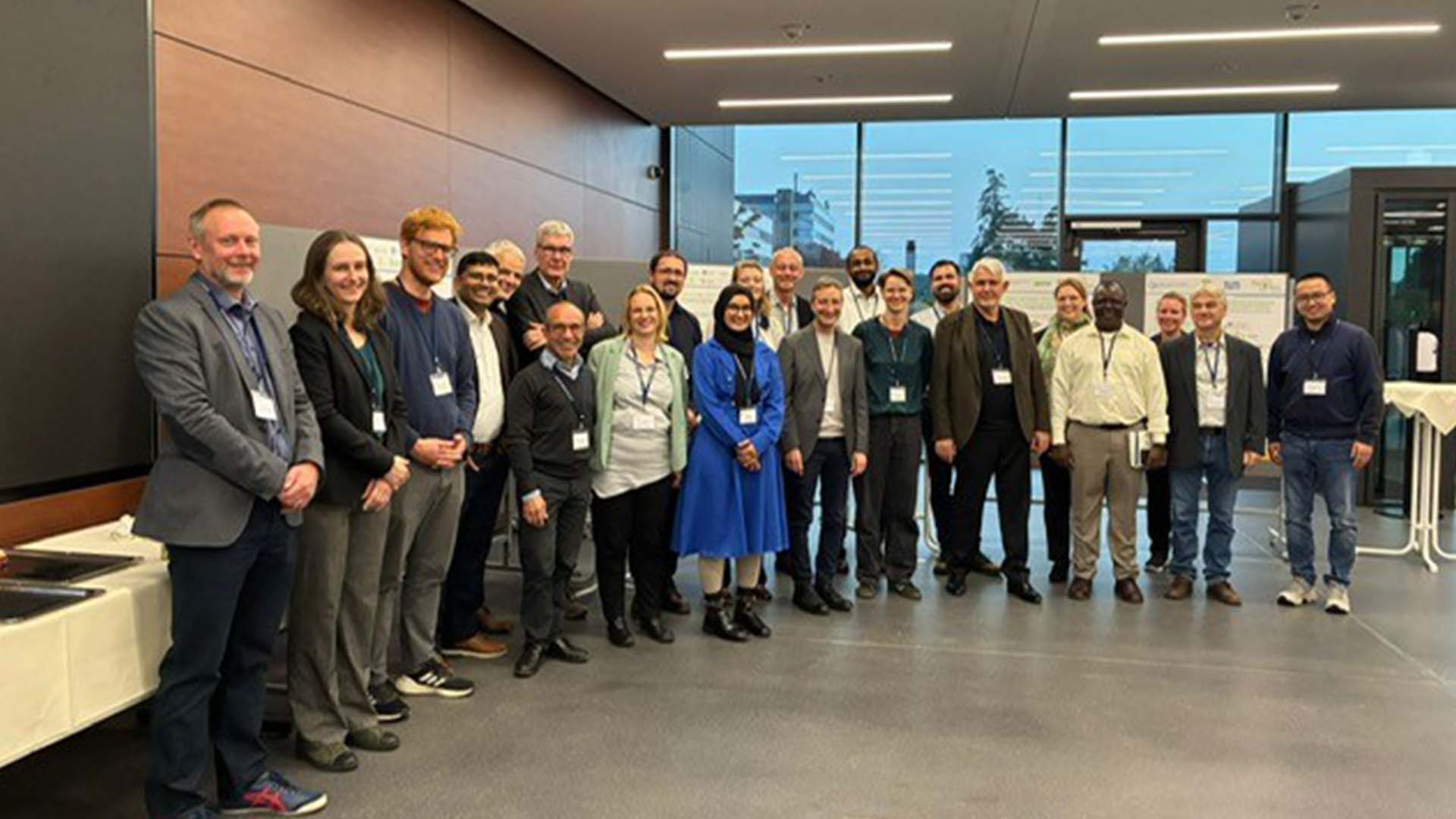 A group of people stands indoors in a conference room, posing for a group photo. Most are wearing business casual attire and name badges, gathering for a MultiStress Research meeting focused on ecophysiology and maize.