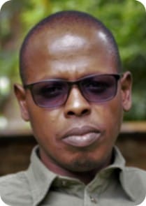 A person wearing dark sunglasses and a green collared shirt looks directly at the camera with a neutral expression. Greenery, hinting at climate-resilient agriculture or maize fields, is visible in the blurred background.