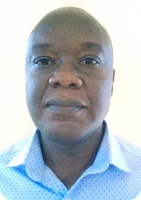 A man with a shaven head wearing a light blue collared shirt looks directly at the camera against a plain, light background, embodying expertise in climate-resilient agriculture.