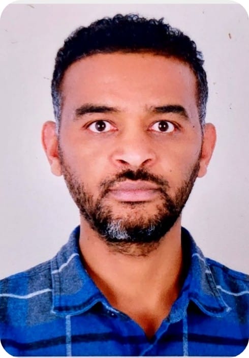 A man with short curly hair and a trimmed beard, wearing a blue checked shirt, poses for a portrait against a light background, representing his work in MultiStress Research to advance crop modelling and promote food security.