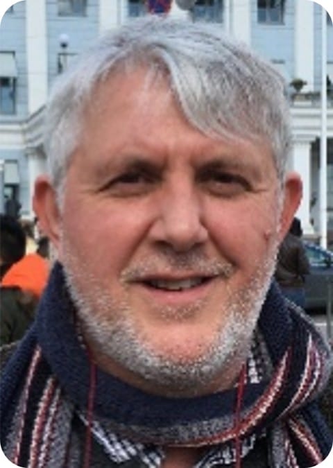 A middle-aged man with grey hair and beard, wearing a scarf and checked shirt, stands outdoors in front of a white building, reflecting on crop modelling techniques for climate-resilient agriculture.
