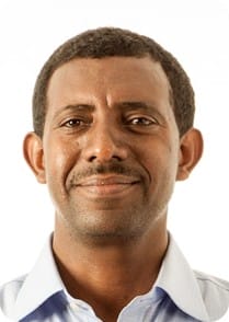 A man with short curly hair, medium brown skin, and a trimmed moustache, wearing a light blue collared shirt, smiles slightly against a white background—reflecting his passion for MultiStress Research in crop modelling.