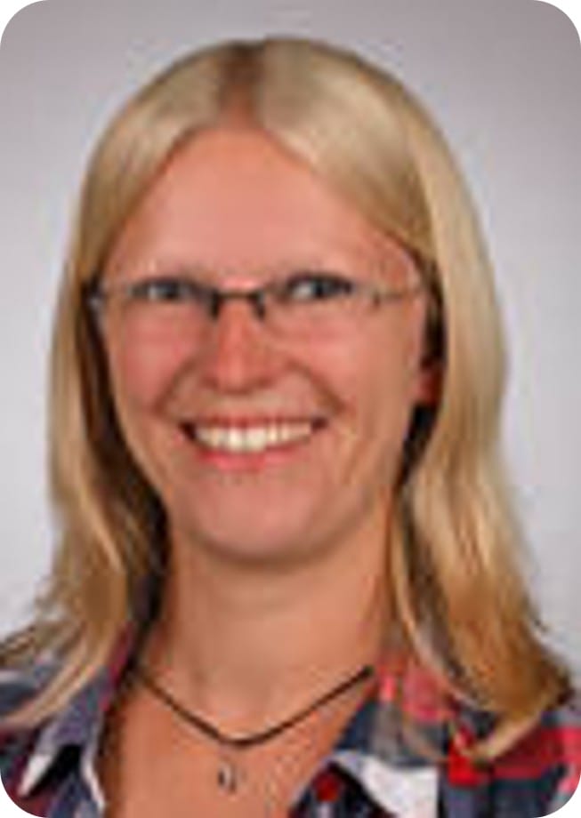 A woman with straight, blonde hair and glasses smiles at the camera. She is wearing a checked shirt and a necklace—perfectly embodying the spirit of climate-resilient agriculture with her dedication to sustainable food security.