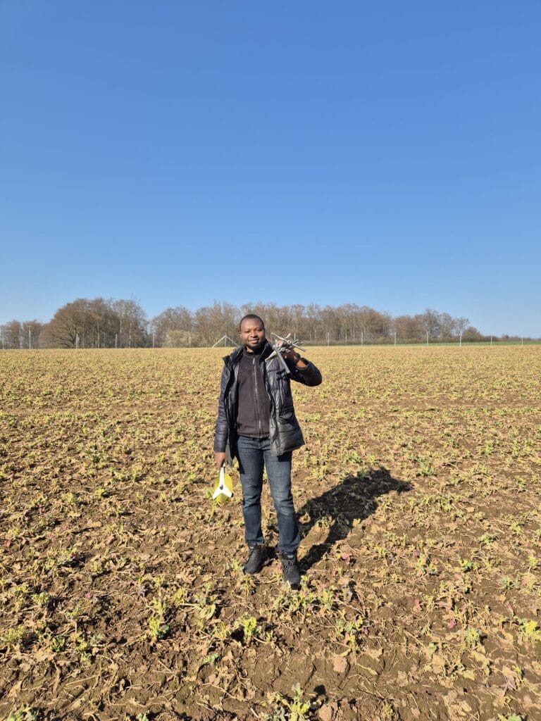 A person stands in a partially planted field holding tools, embodying climate-resilient agriculture, with trees and a clear blue sky in the background.