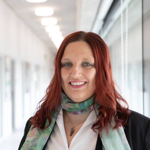 A woman with long red hair, wearing a patterned scarf and black jacket, stands smiling in a brightly lit corridor with windows, reflecting her commitment to advancing food security and climate-resilient agriculture.