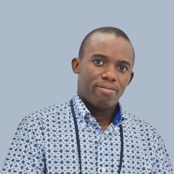 A person wearing a light blue patterned shirt and a lanyard stands against a plain light blue background, representing Climate-Resilient Agriculture and innovative Multi-Stress Research in the field.
