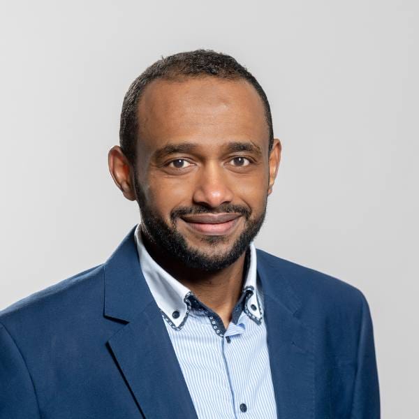 A man with short hair and a beard, wearing a blue suit jacket over a light blue collared shirt, smiles at the camera against a plain light grey background, reflecting his expertise in maize ecophysiology and crop modelling.