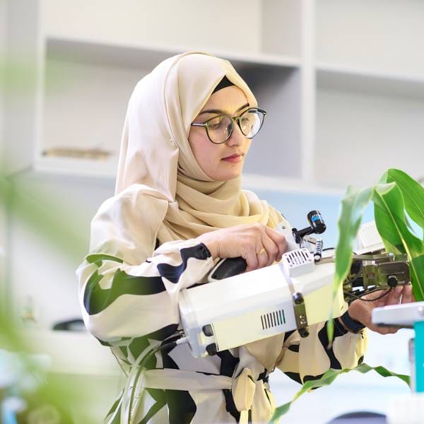 A woman in a hijab uses scientific equipment to examine a green plant in a laboratory, advancing MultiStress Research to support food security.