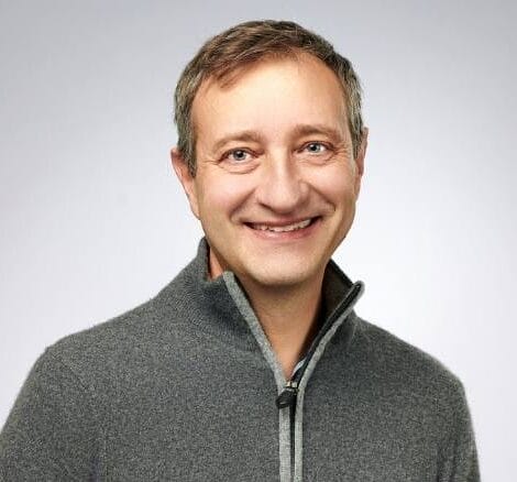 A man with short brown hair smiles at the camera, wearing a grey zip-up jumper against a plain light grey background, reflecting his expertise in MultiStress Research and climate-resilient agriculture.