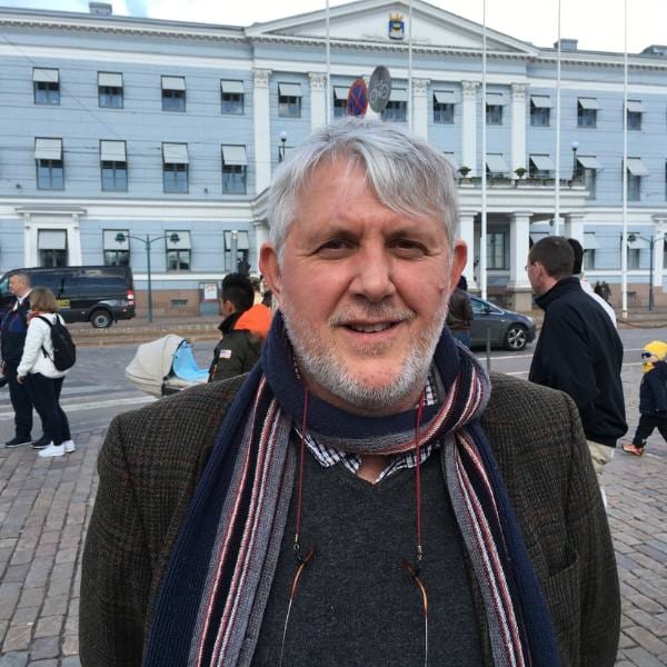 A man with grey hair and a beard stands outdoors wearing a scarf and jacket. Several people and a large blue building, possibly related to maize research or food security, are visible in the background.