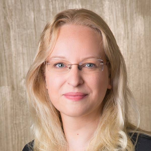 A person with long blonde hair and glasses smiles softly, wearing a black top, in front of a light brown textured background, reflecting on ecophysiology research for maize and its impact on food security.