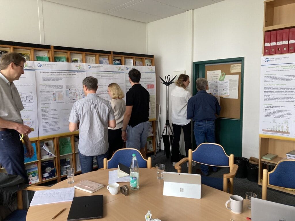Six people stand and review posters and papers on MultiStress Research in ecophysiology, displayed on a wall in an office meeting room with bookshelves, chairs, and a table with drinks and devices in the foreground.