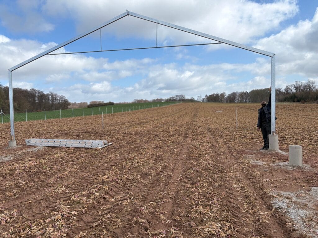 A person stands next to a metal frame structure on a ploughed field under a cloudy sky, demonstrating climate-resilient agriculture practices amid open farmland and trees in the background.