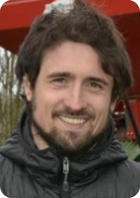 A man with dark hair and a beard smiles at the camera, wearing a black jacket. The background includes trees and part of a red structure, suggesting he might be involved in maize crop modelling or related ecophysiology research outdoors.
