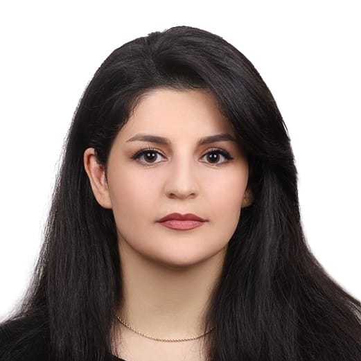 A woman with long dark hair and neutral make-up is facing the camera against a plain white background. She is wearing a black top and a thin gold necklace.