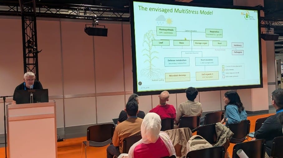 A presenter speaks at a lectern in front of an audience, with a slide titled "The envisaged MultiStress Model" displayed on a large screen.