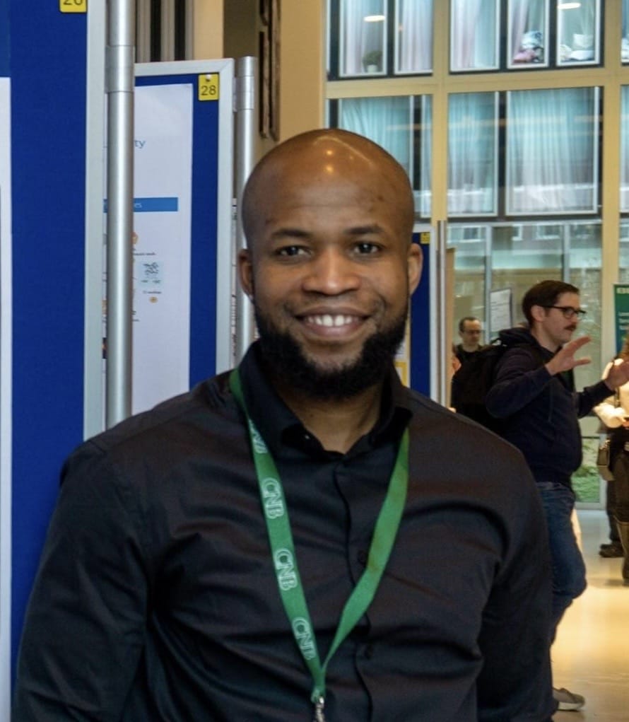 A man wearing a black shirt and green lanyard stands indoors, smiling at the camera; people and posters are visible in the background.