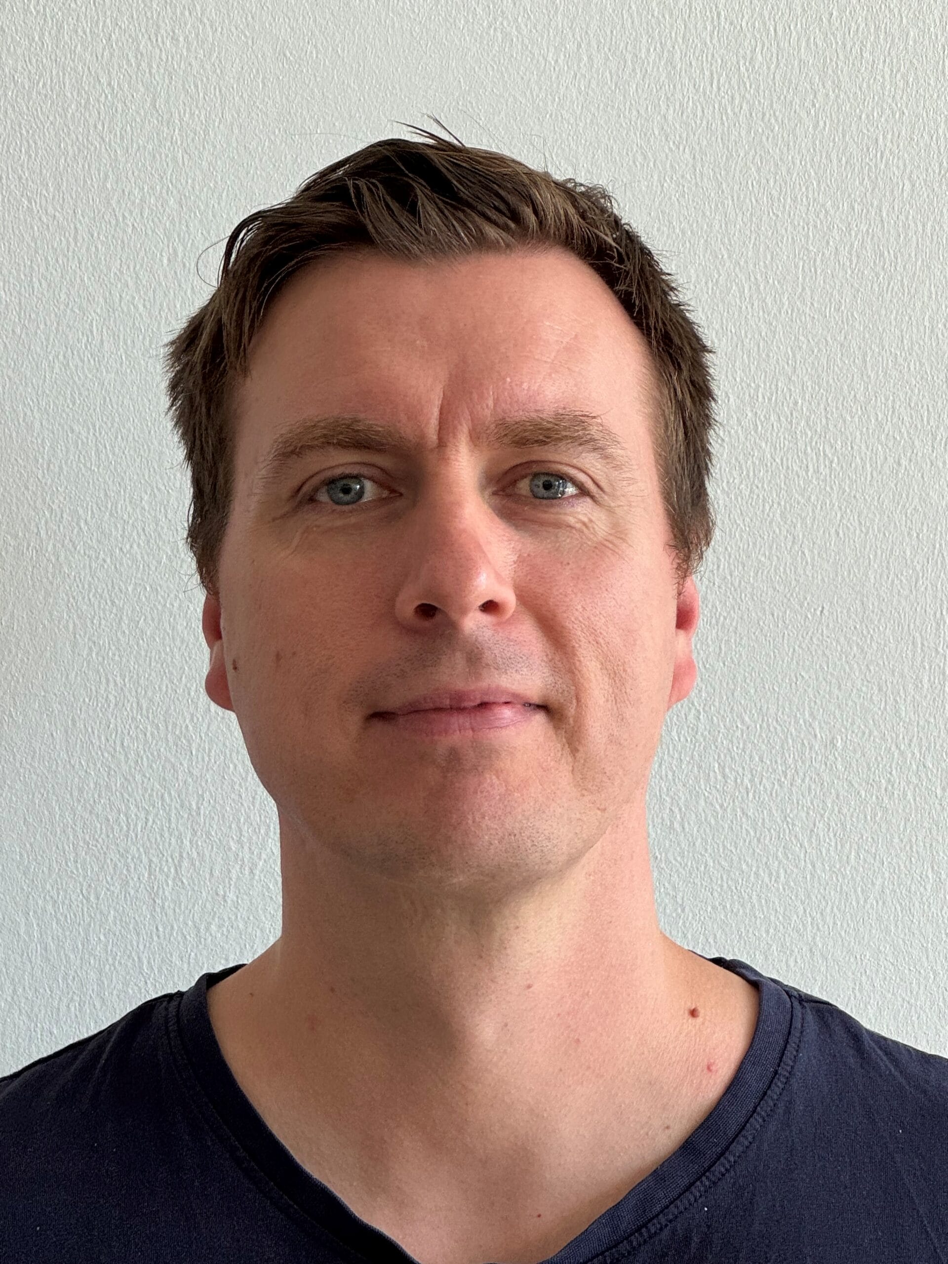 A man with short brown hair and blue eyes is facing the camera against a plain white background, wearing a dark crew-neck jumper.