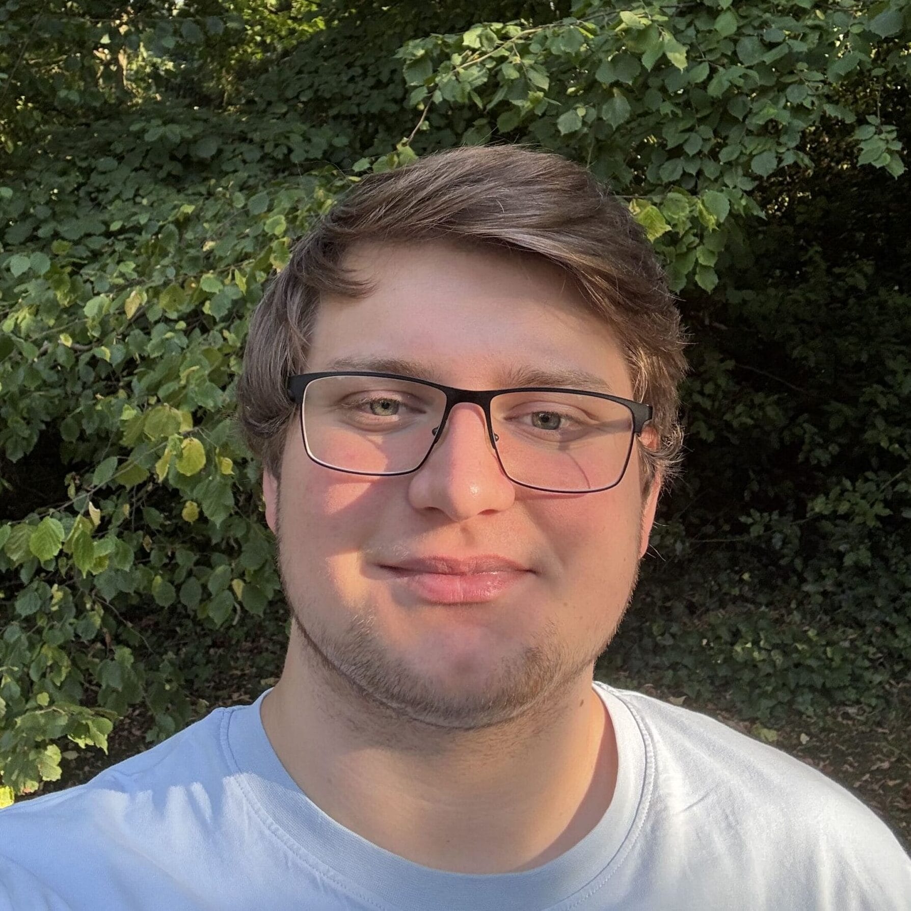 A person with glasses and short brown hair takes a selfie outdoors, with green trees and sunlight in the background.