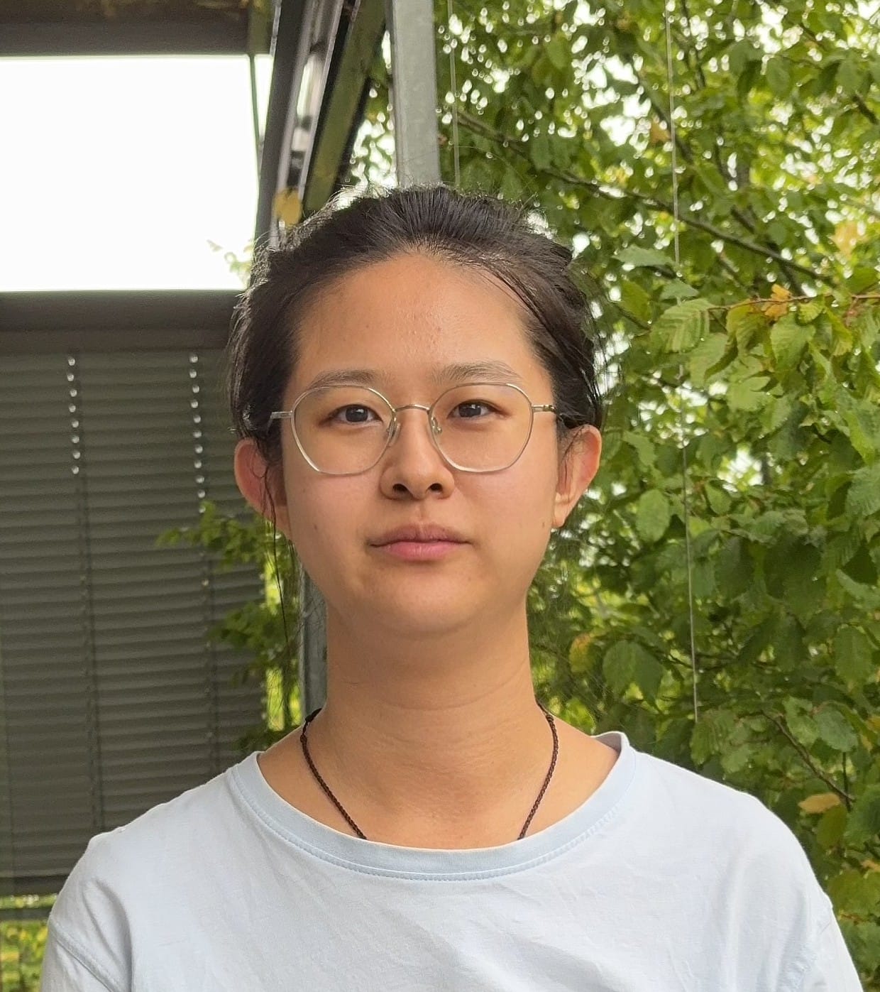 Person with glasses and tied-back hair stands outside, wearing a light-coloured shirt, with greenery and a metal structure in the background.