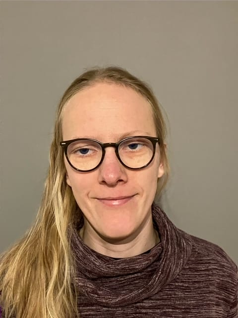 A person with long blonde hair and glasses, wearing a maroon jumper, stands in front of a plain grey background.